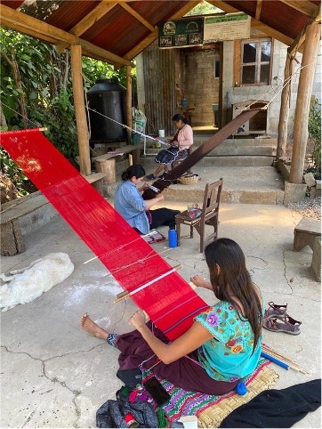 Three people sitting in a covered, outdoor area holding and working on large weavings