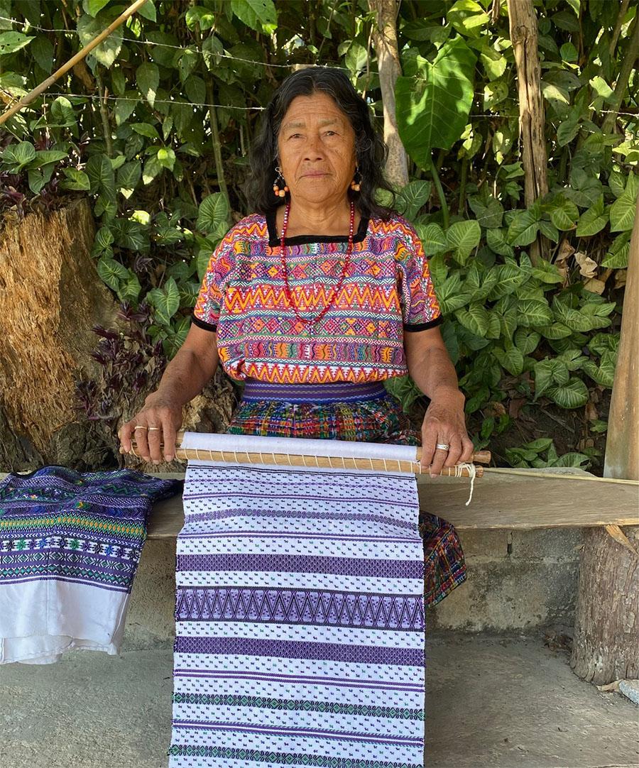 A seated woman in a colorful shirt and skirt holding a large, woven piece of fabric