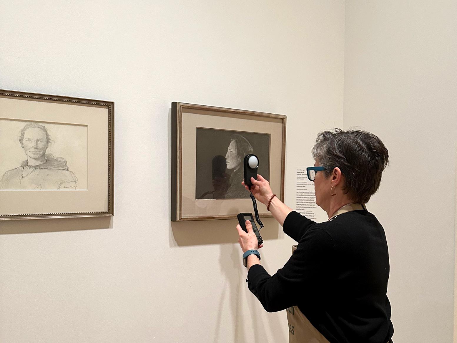 A woman holds a light meter in front of an artwork on paper hung on a gallery wall