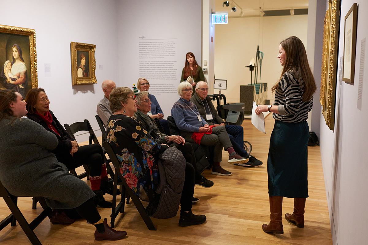 A person standing along a wall hung with paintings and speaking to a seated group of older adults