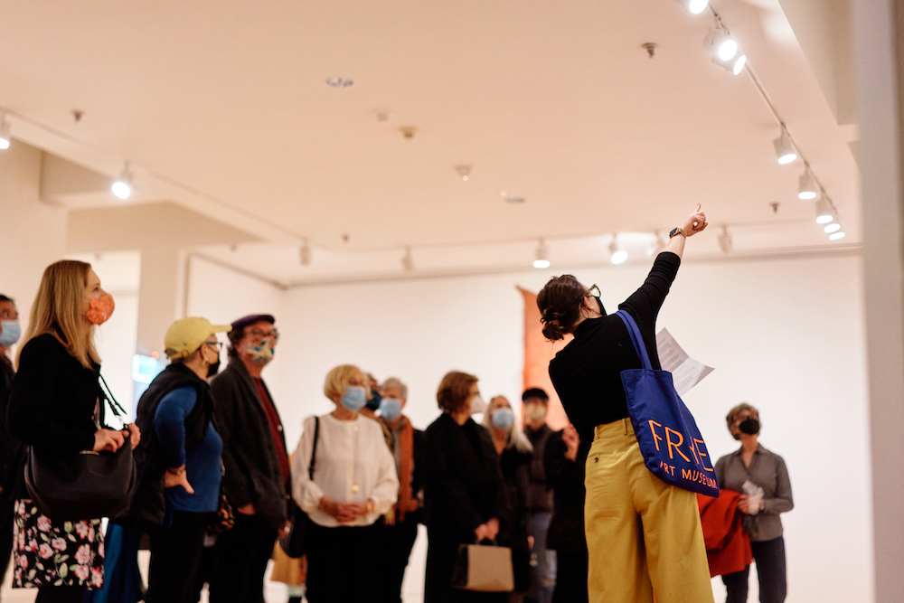 A tour guide points up at an unseen artwork while a masked tour group looks on