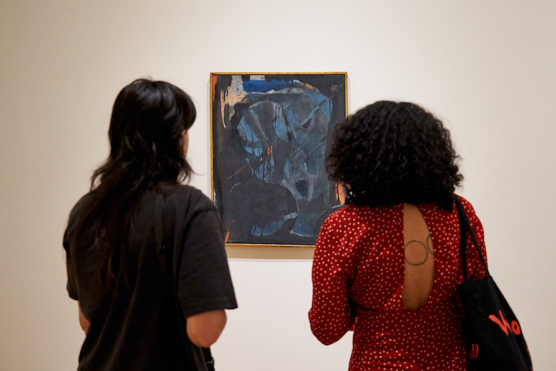 The back of two visitors observing a painting on a gallery wall