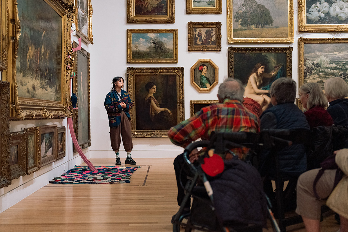 A person standing next to walls covered in paintings and talking to a seated group of older adults