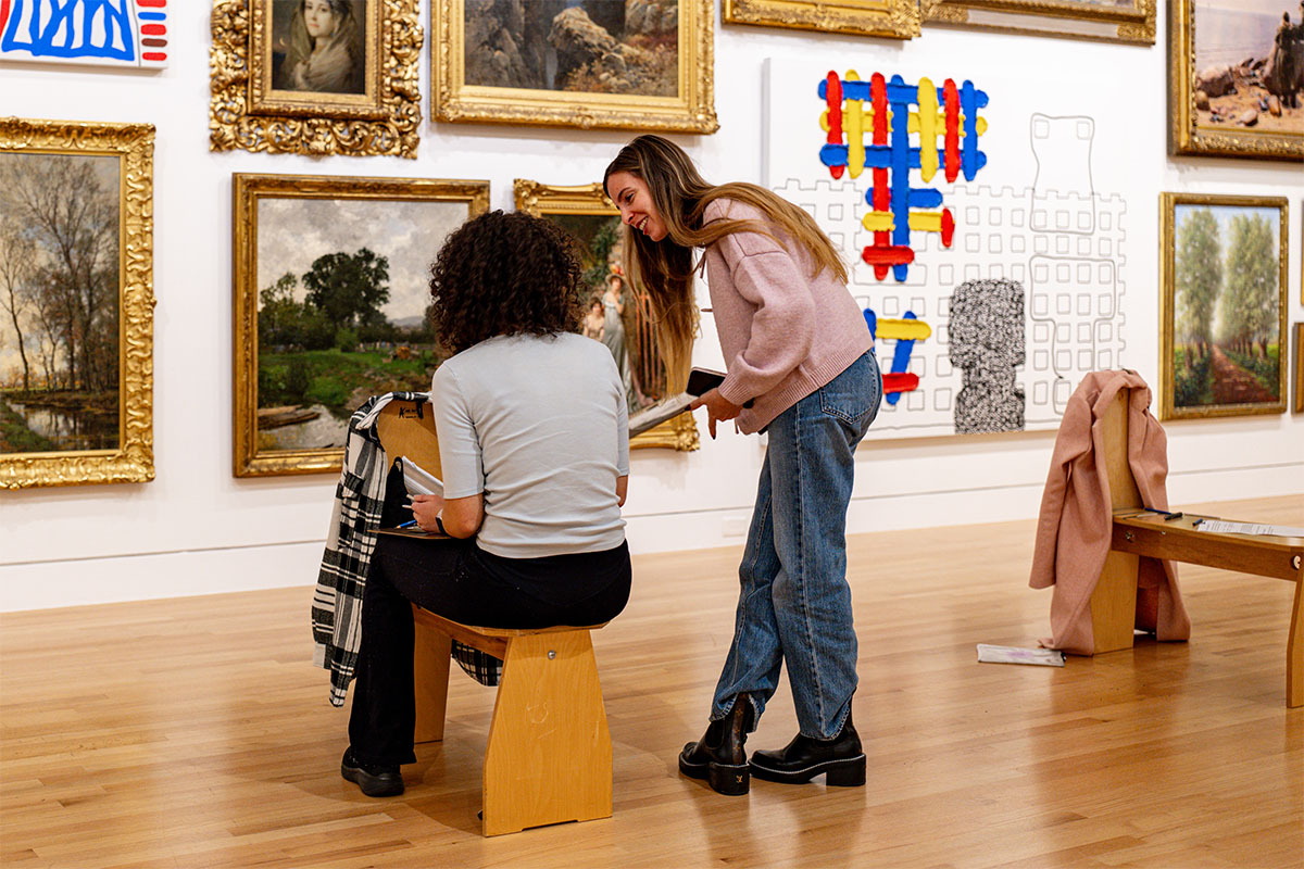 Two people chatting while holding clipboards in front of a large wall covered in paintings. One is sitting on a drawing stool and the other is standing nearby.