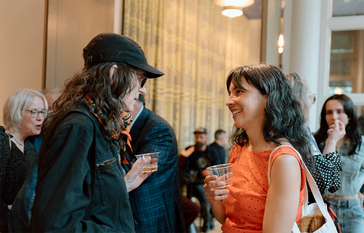 Two women in profile chatting while one holds a disposible cup of white wine in a well lit reception area