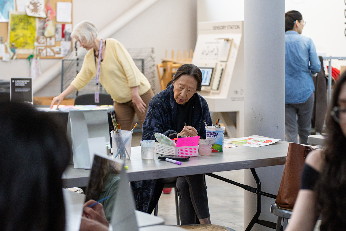 A person sitting and working on an art project. Other people are visible doing the same in other pages of the room.