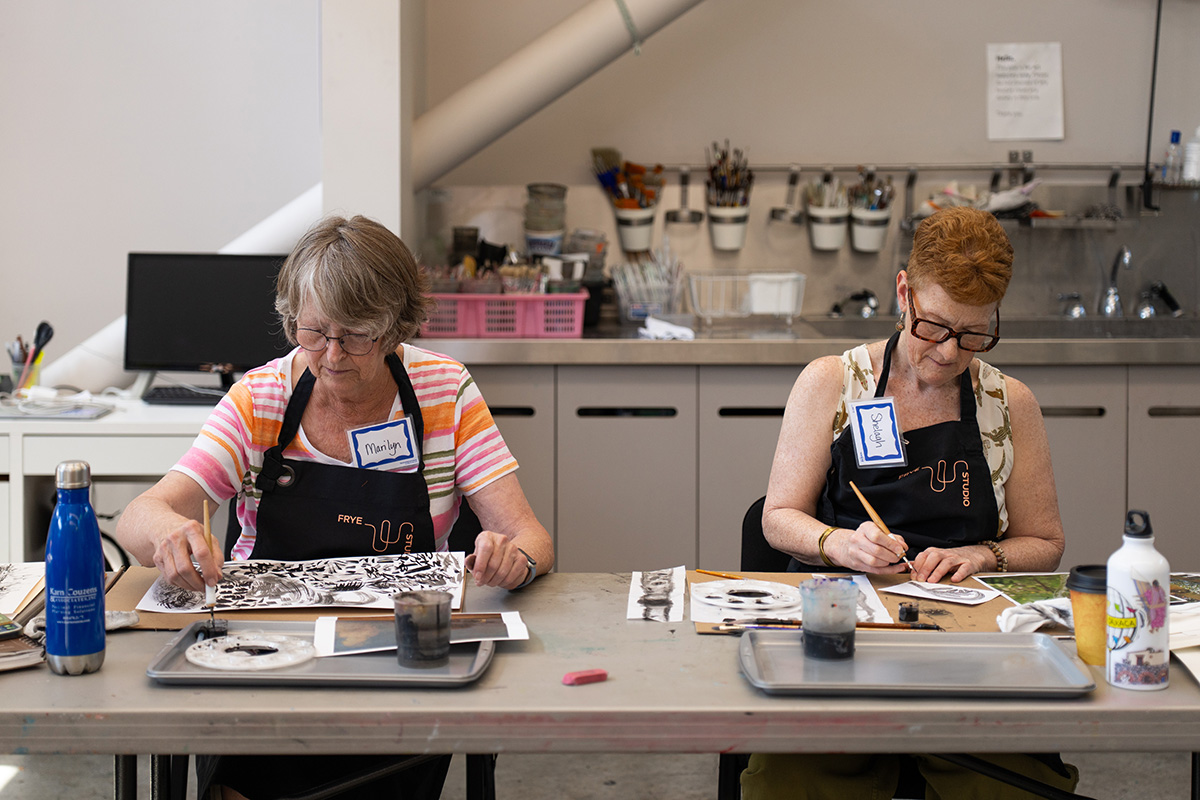 Two older people sitting side by side at a long table and working on art projects in a studio space