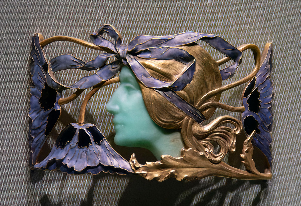 A wide, elaborate brooch with a green face in profile in the center and a surrounding insect wings motif.
