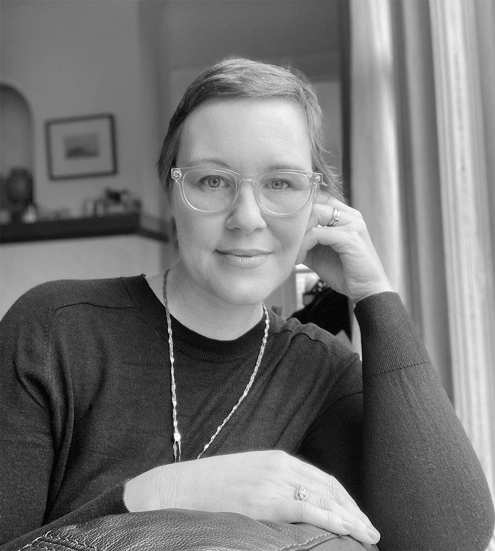 A black and white photograph of a woman wearing glasses and short hair leaning against the back of a seat while looking at the camera