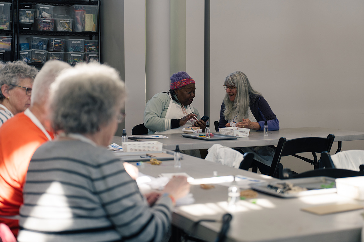 Older adults seated at long tables and chatting in an art studio space