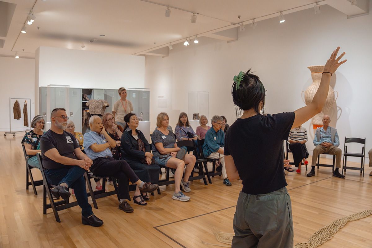 A group of seated older adults in an art gallery space being spoken to by a person whose back is to the viewer