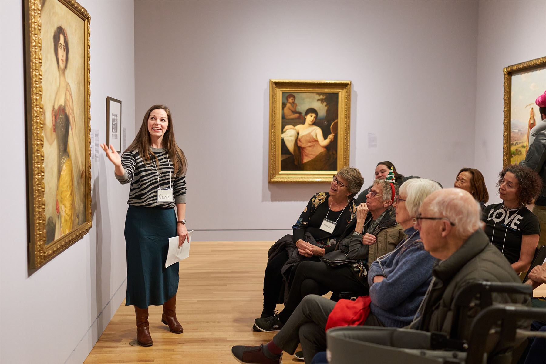 A women standing in front of a seated group and gesturing at a painting on the wall