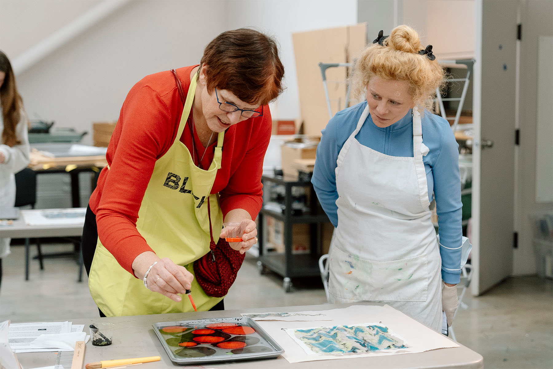 Two women engaged in an art activity. They are wearing aprons and looking at a tray with liquid and spots of color on a table in front of them.