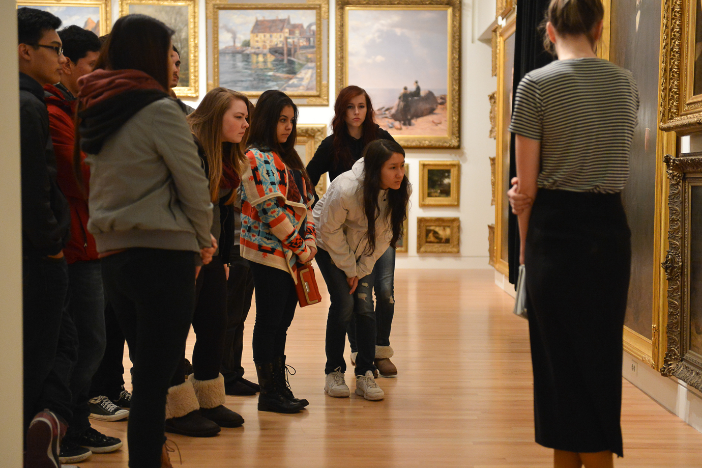 Photo of a group of teenagers looking closely at a painting on a gallery wall with paintings hung in a salon style behind them