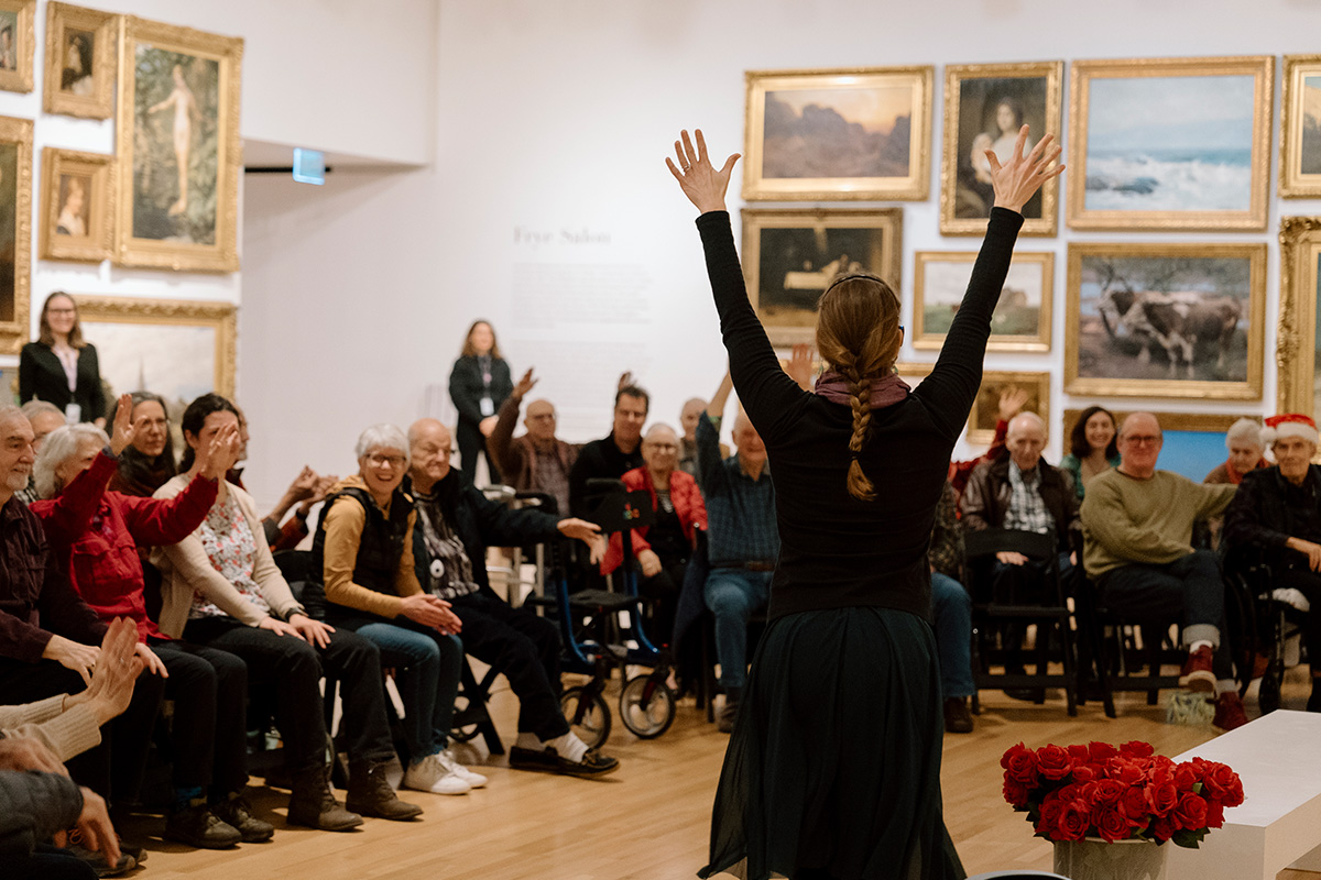 A group of older adults being led in a seated dance activity in a large gallery space