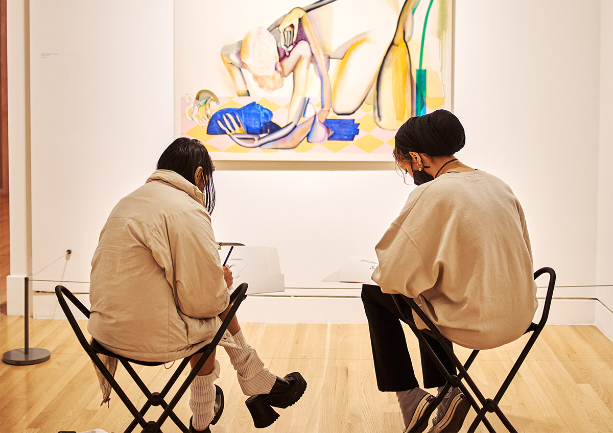 Two younger people sitting on stools and drawing on notepads held in their laps in front of a large painting