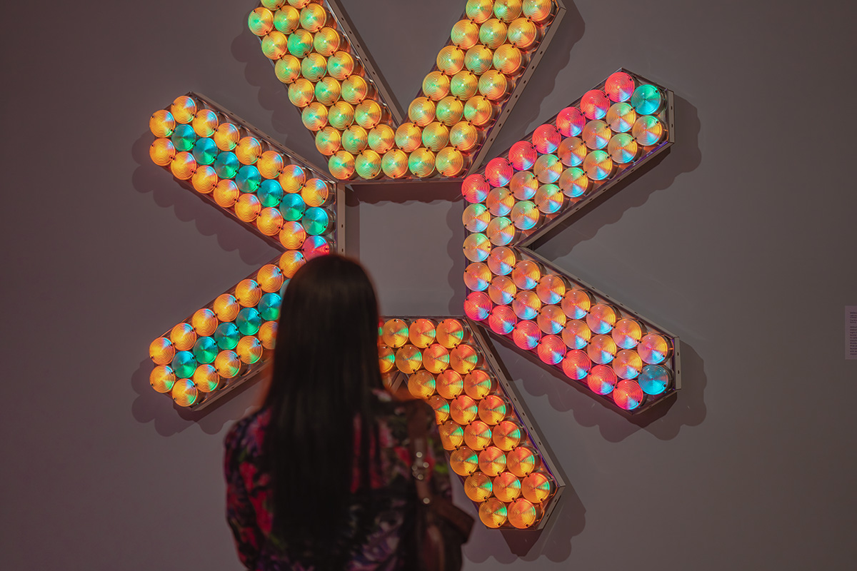 A person viewed from behind standing in front of a large, wall mounted artwork made out of many colorful lights