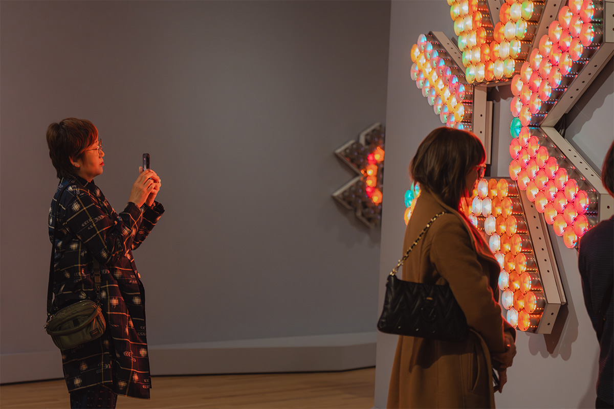 People standing in front of a large, geometric, wall mounted sculpture made up of a number of lightbulbs in different colors. One person is taking a photo of the work on their phone.