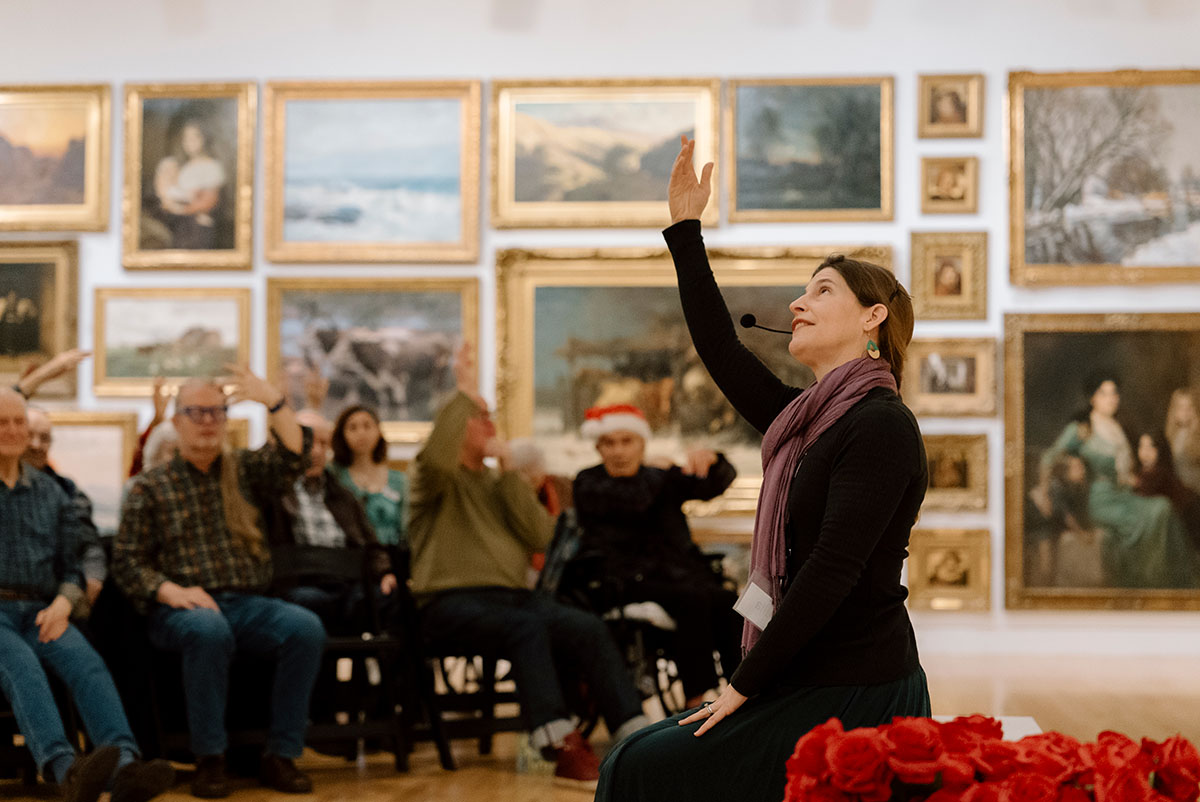 A person in profile sitting on a bench with a hand raised speaking to a group of people in a room with many paintings on the walls.