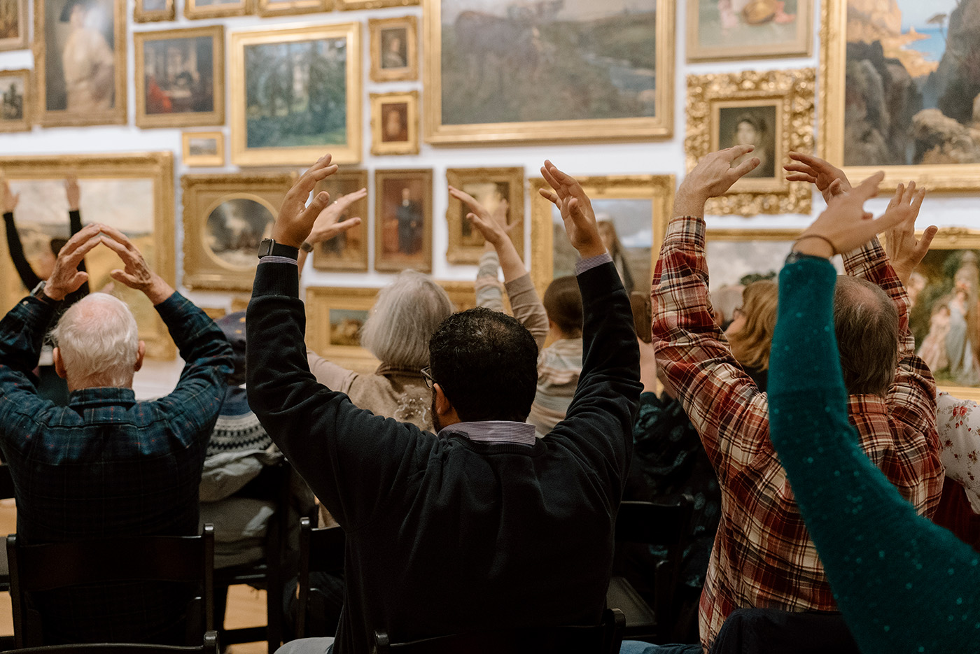 Seated people viewed from behind holding their hands above their heads in an O shape during a moment exercise in an art gallery
