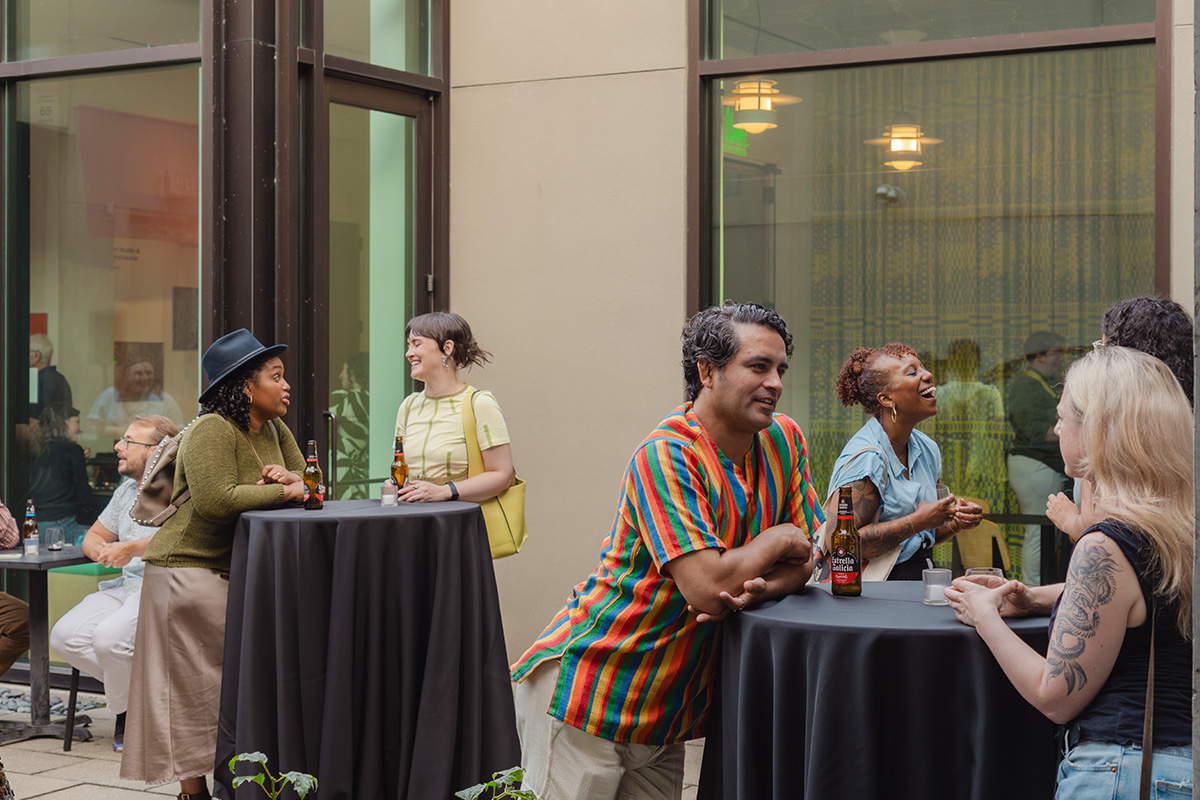 People in an outdoor courtyard in the evening standing at tall tables with drinks and chatting