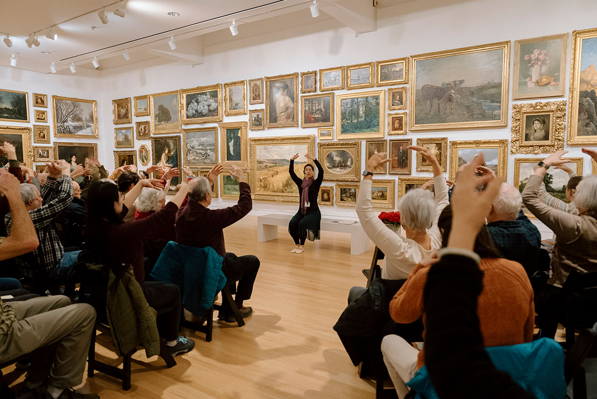 A person seated in front of a wall covered in paintings and facing a semi-circular group of people raises their hands over their head.