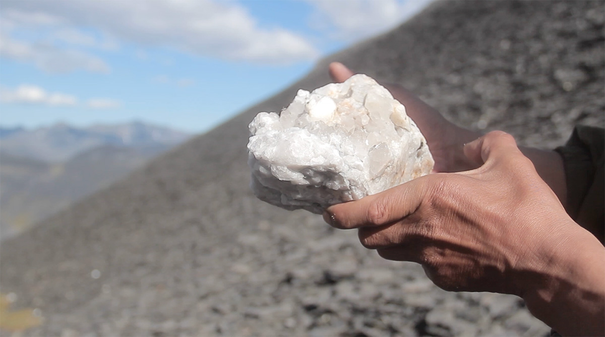 A still from a video of a close up of hands holding a large white rock in a rocky mountain landscape