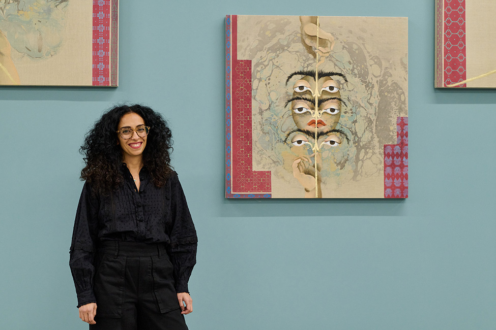 A woman wearing black with curly black hair smiling and standing in front of a blue wall hung with multiple paintings