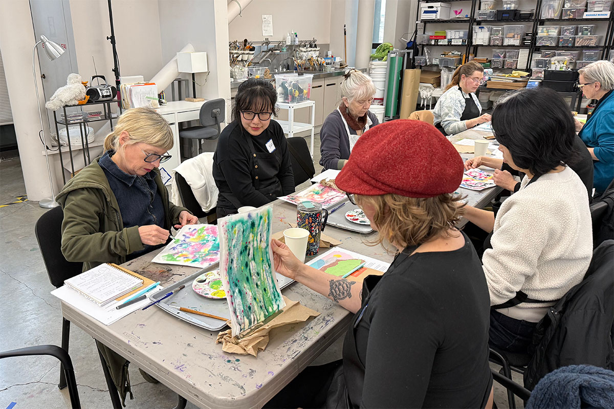 People sitting at a long table in an art studio space working on colorful paintings