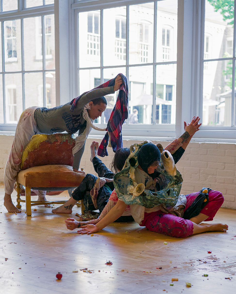 Dancers covered in sculptures and paint performing around a chair