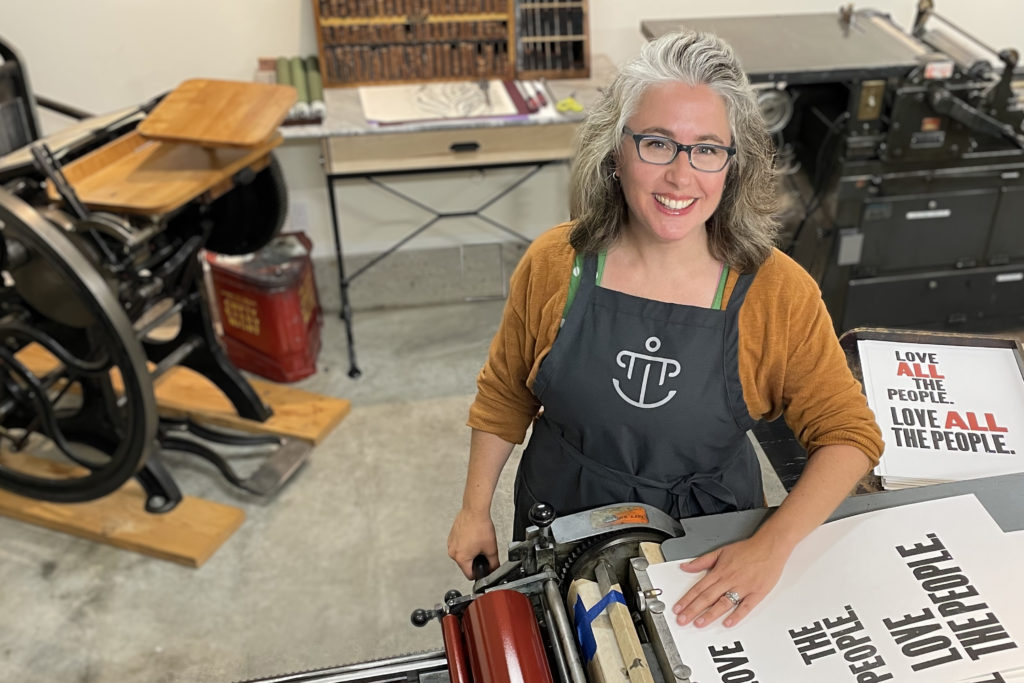 A photograph of a woman wearing an apron standing in front of a printing press