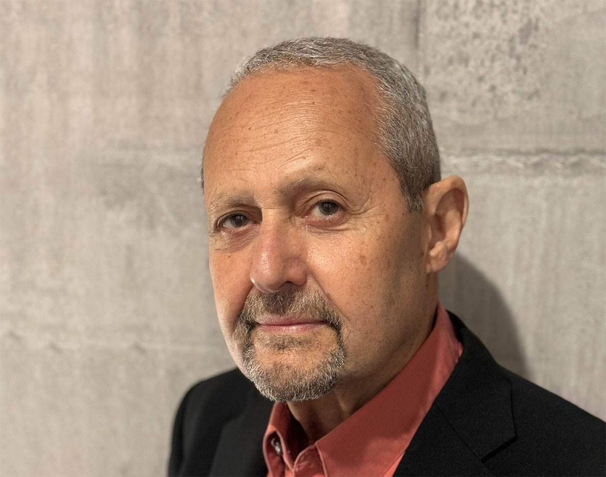 A slightly angled photo of a man in a suit standing in front of a wall