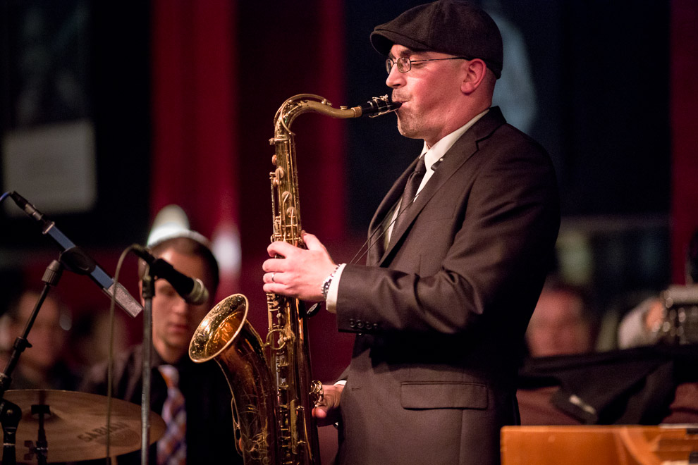 Photograph of Kareem Kandi playing a saxophone on stage with other musicians behind him