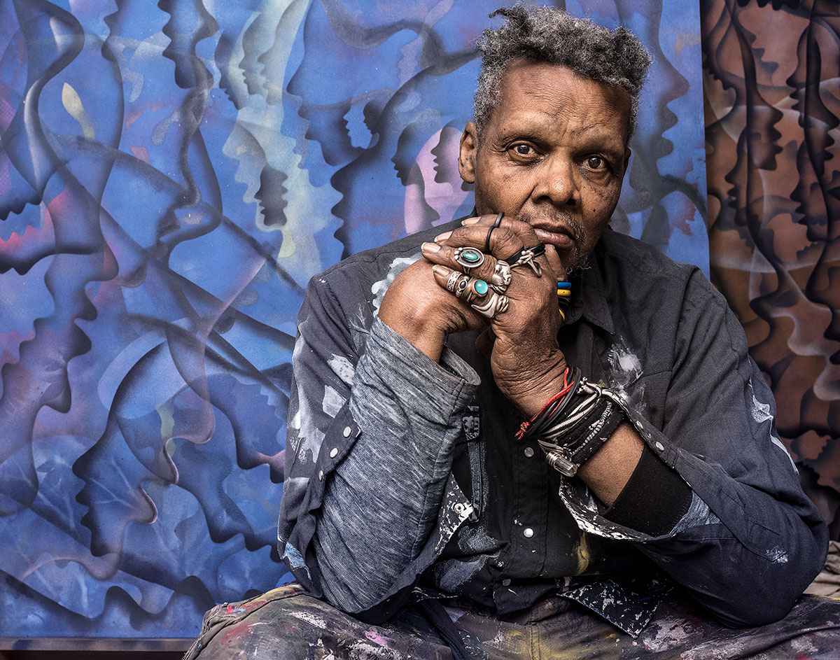 An older black man leaning his elbows on his knees and wearing clothes splattered with paint in front of a large painting