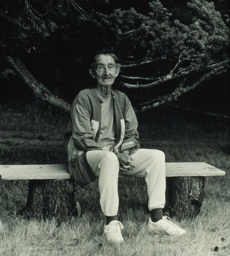 A black and white photograph of an older woman sitting on an outdoor bench
