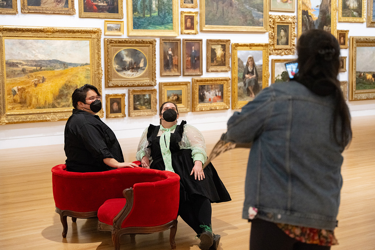 Two people wearing masks posing for a casual photo in a circular red sofa chair in a large room with paintings on the walls