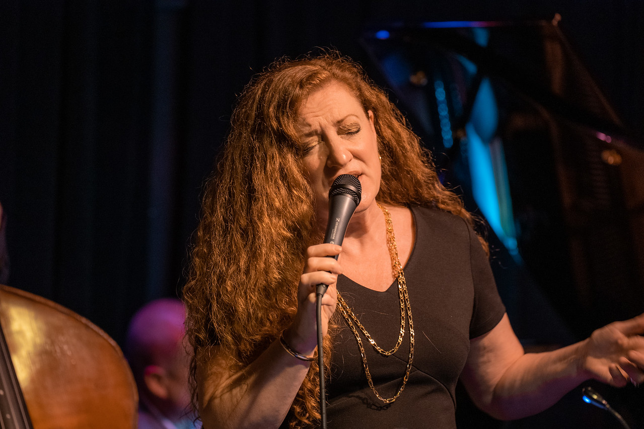 A woman with long, red curly hair singing into a microphone on a stage