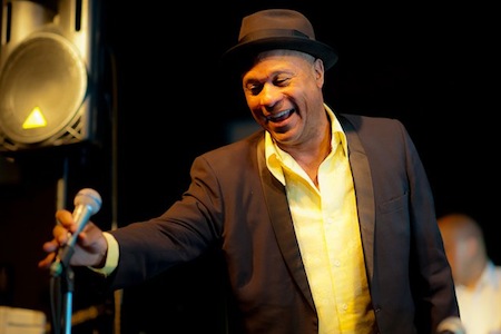 Photo of Reggie Goings, a man wearing a suit jacket, yellow shirt, and a fedora holding a microphone on a mic stand and smiling