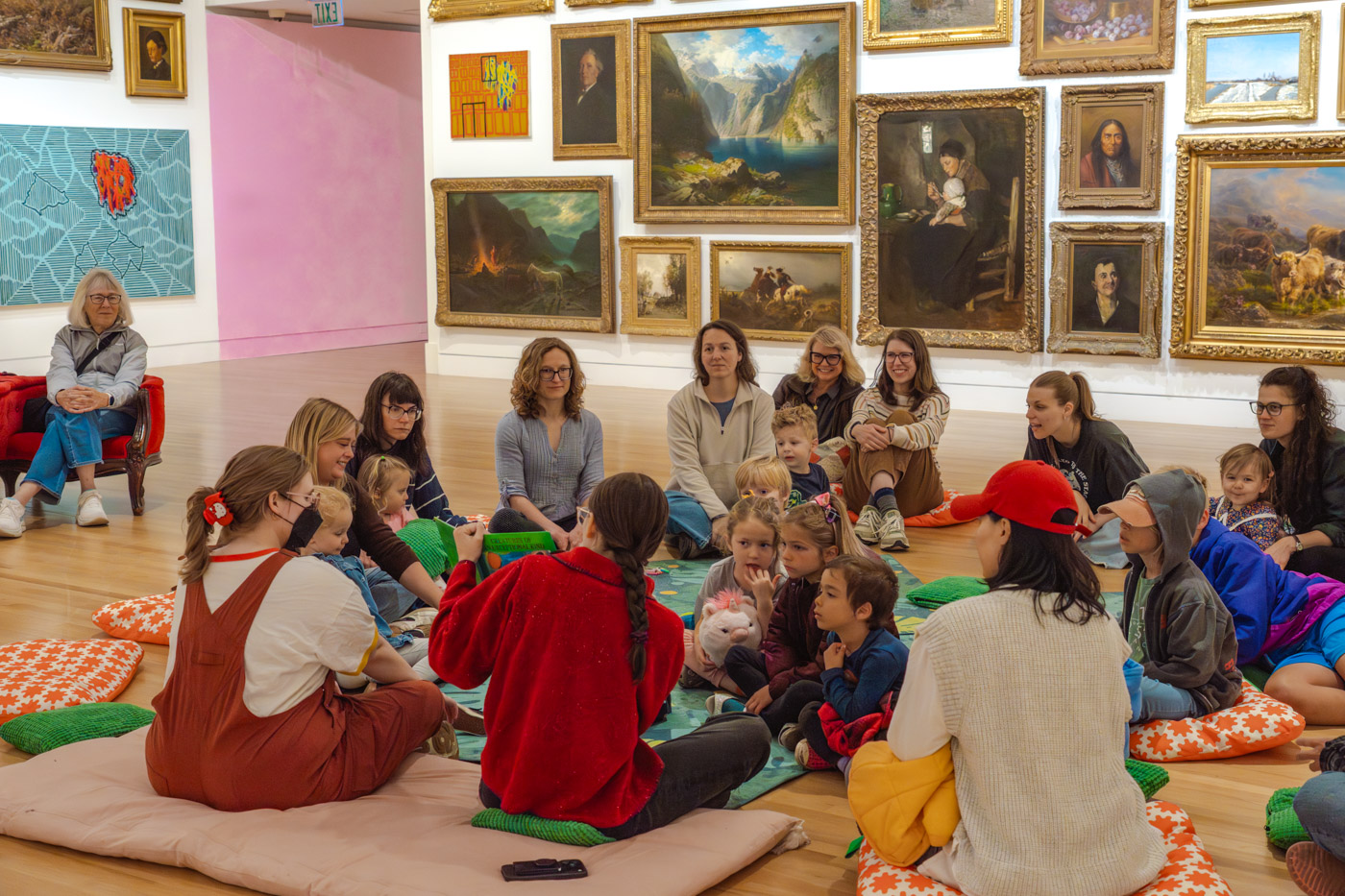 A group of adults and children sitting on the floor of a large gallery space watching someone read and show image in a picture book