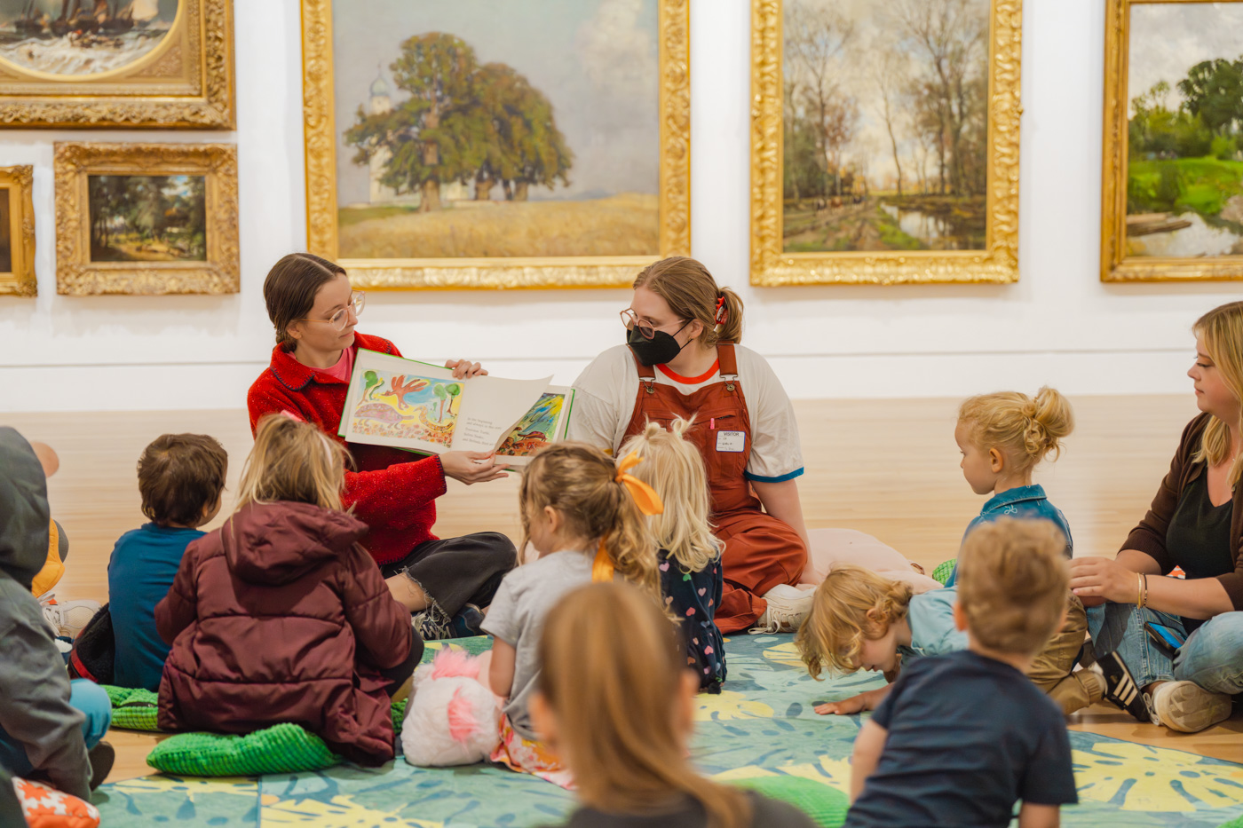 A pair of seated adults read from a children's book to a group of seated adults and chilren in an art gallery space