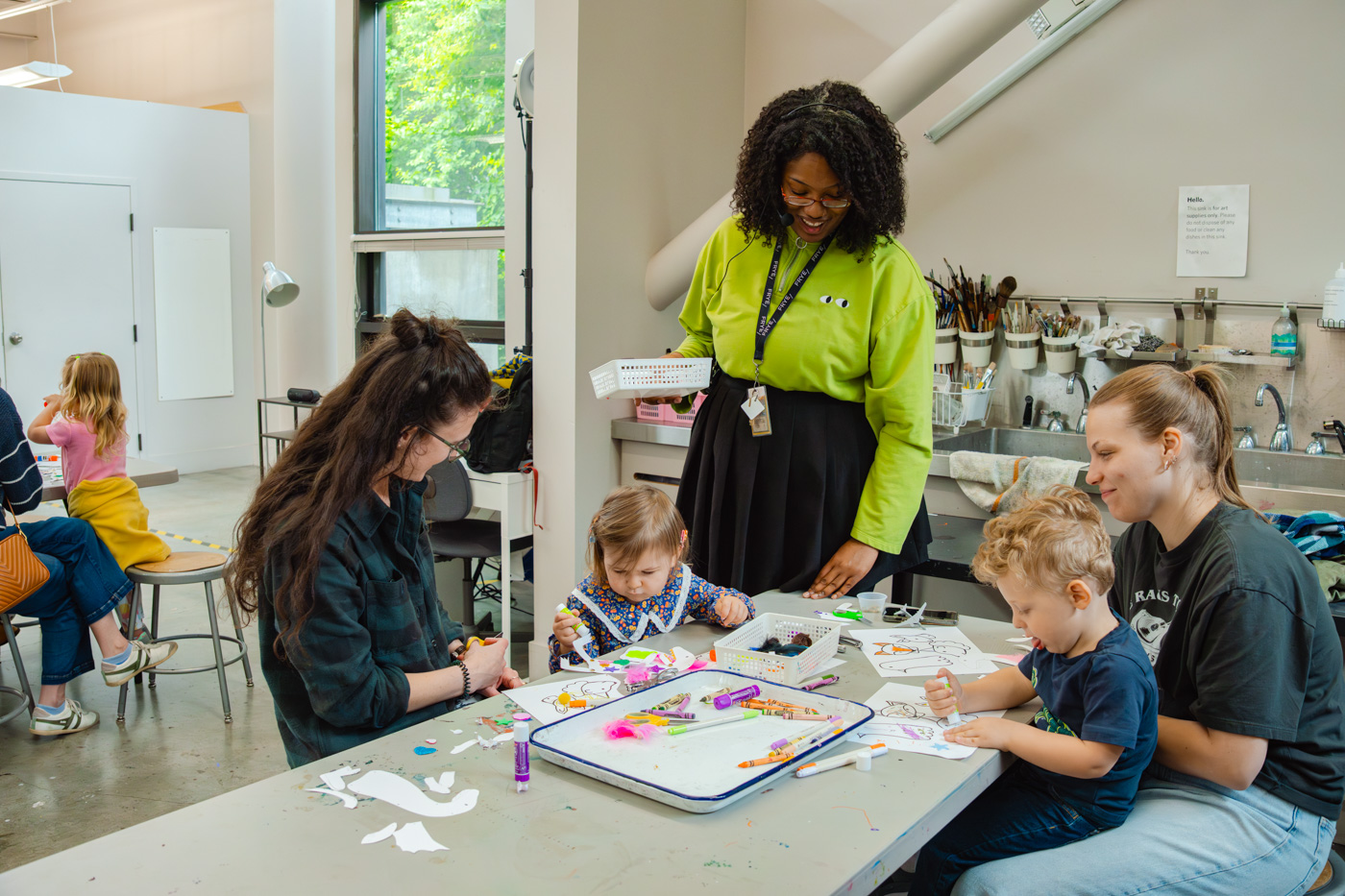 A pair of adults and children working on an art project while a teaching artist standing nearby smiles