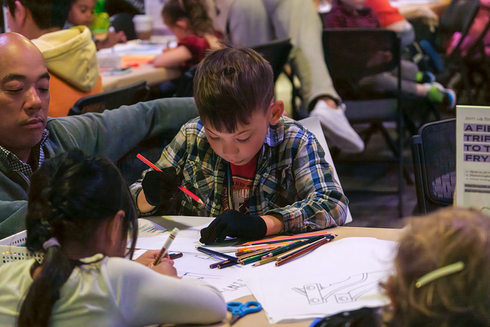 A child drawing with colored pencils on paper at a table with other adults and children