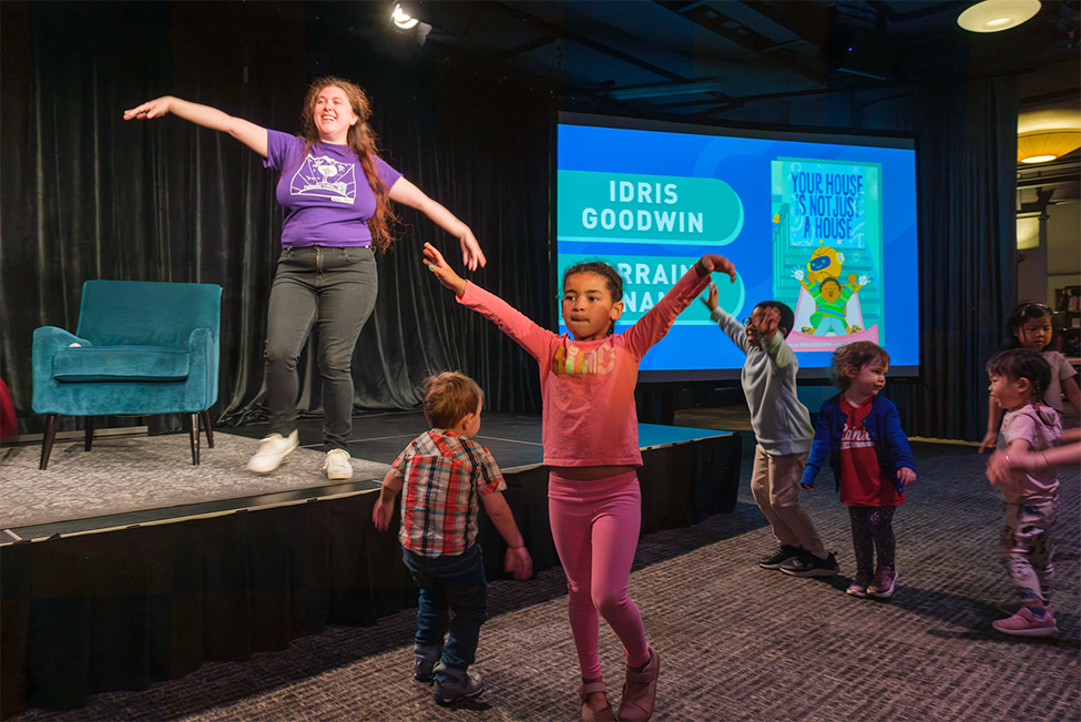 Children standing and engaged in an activity guided by a person raising their arms on a low stage 