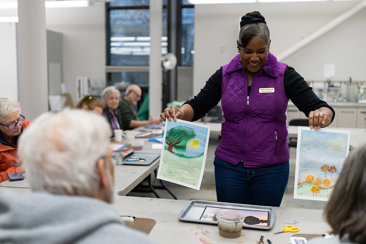 A woman holds up two fresh water color paintings in front of people sitting at a long table