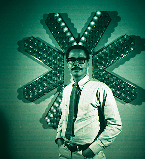 A strongly green-tinged film portrait photo of a man standing in front of a large, geometric, wall-mounted sculpture.