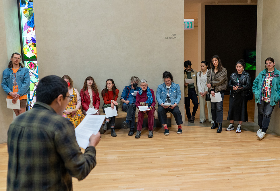 A person with their back to the viewer looking at a paper they're holding and reading aloud to a group of people arrayed along the wall in a circular room