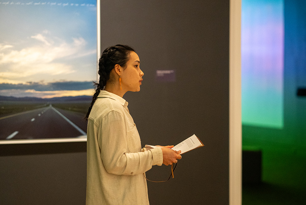 A younger adult woman in profile wearing a white button down and holding a paper speaking in front of a wall-mounted artwork