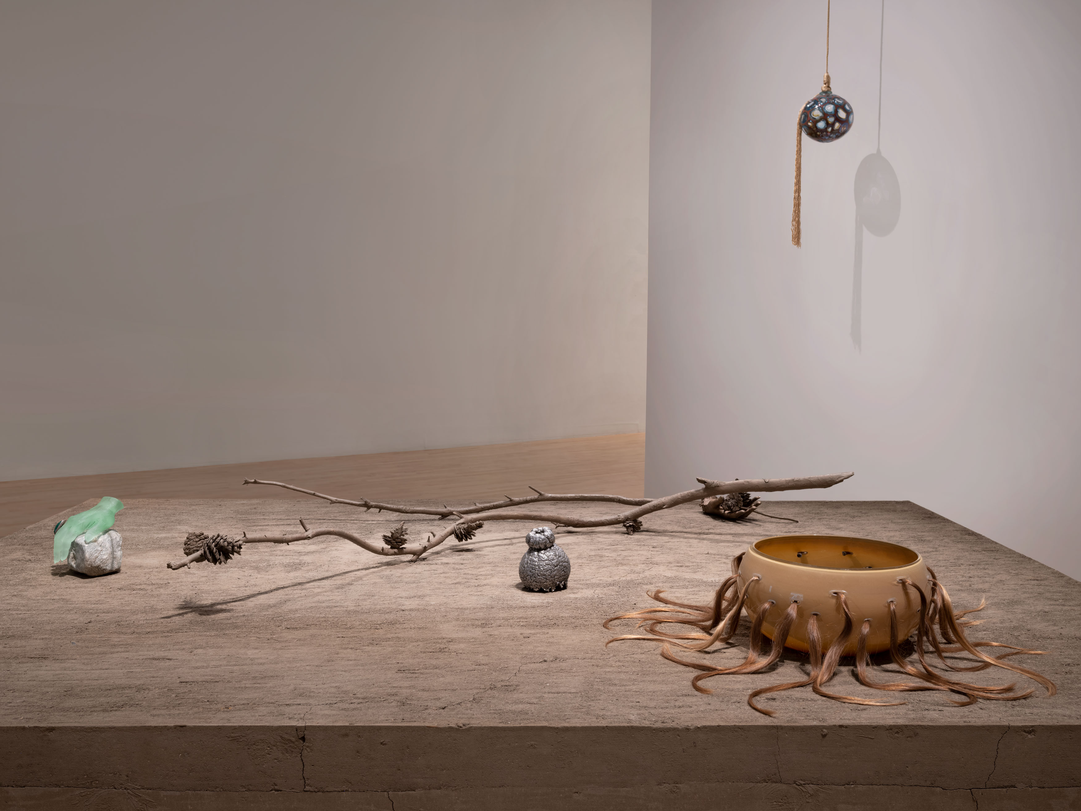 Photo of an exhibition installation with mixed media sculptures displayed on a rammed earth pedestal and a glass sculpture hanging from the ceiling
