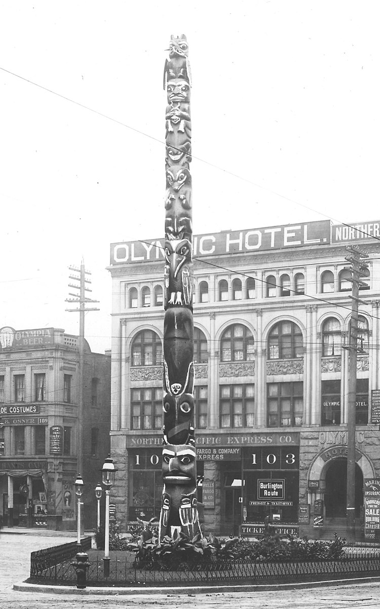 An old black and white photograph of a totem pole in front of a large city building