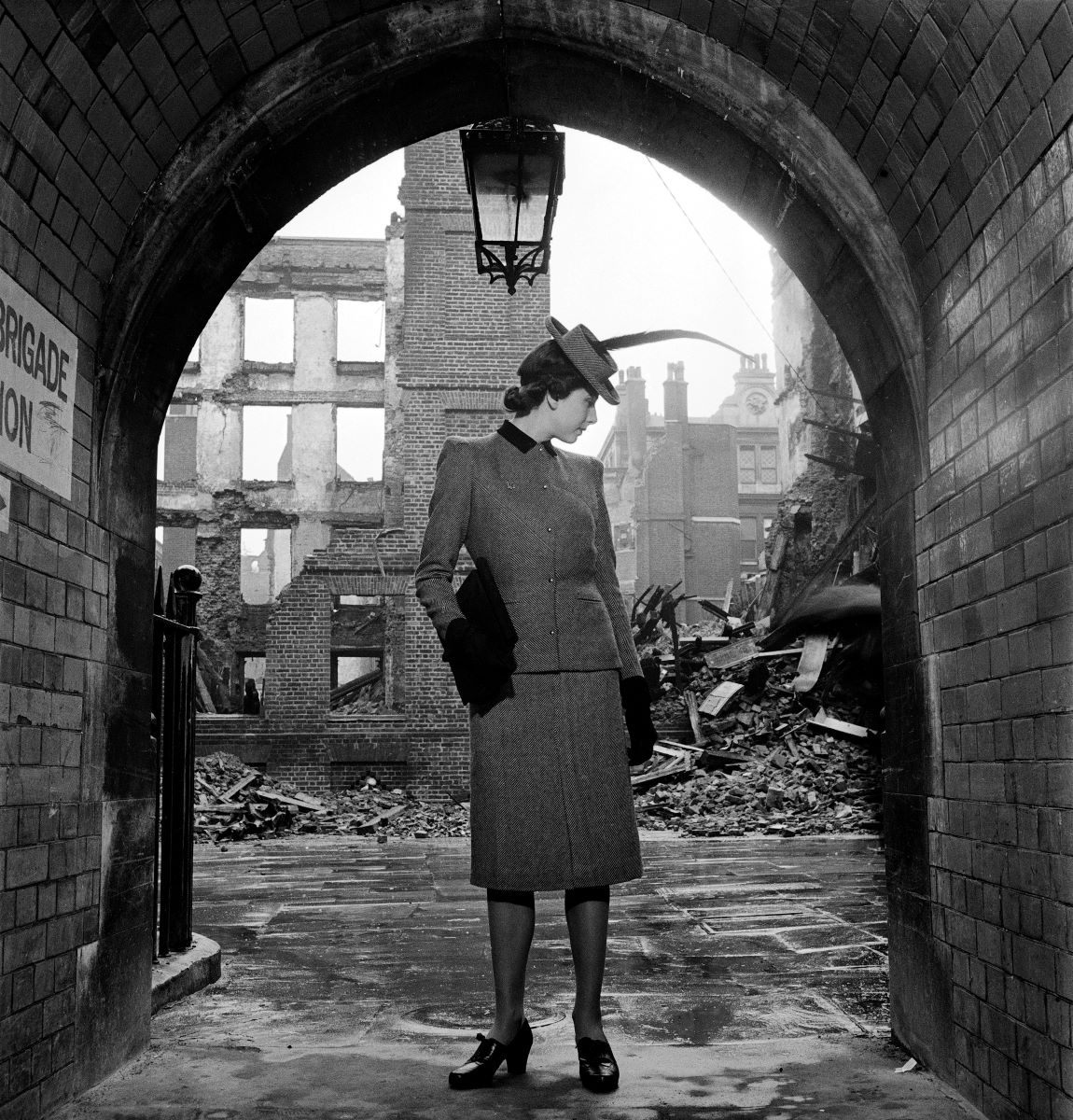 Black and white photo of a woman posing in a long skirt and jacket in front of semi-destroyed buildings 